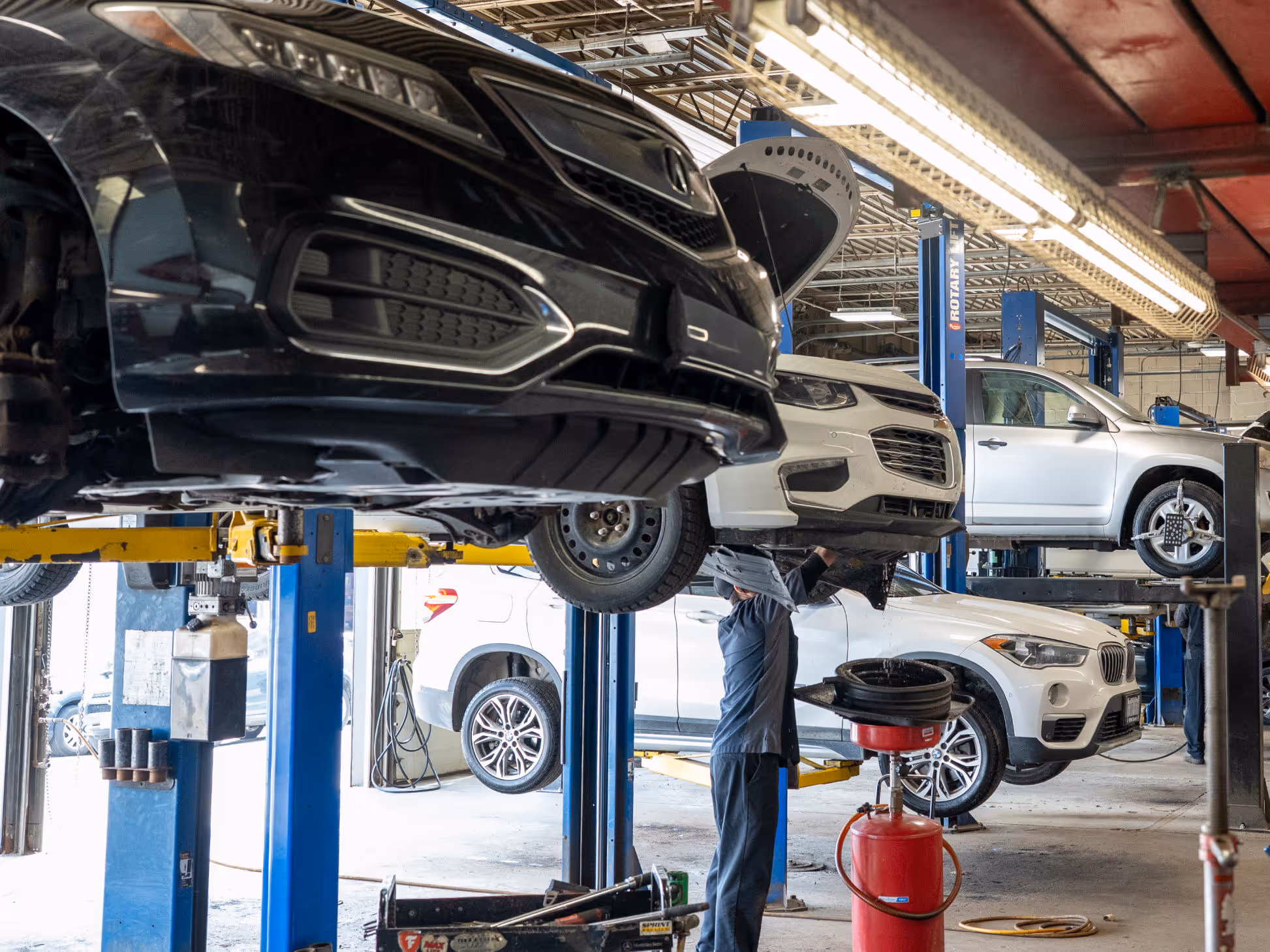 Mechanic working under a white car lifted on a blue hydraulic lift inside an auto repair shop.