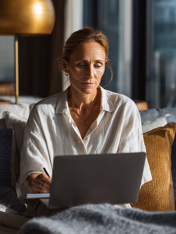 Femme concentrée travaillant sur un ordinateur portable à la maison avec un stylo et un carnet.