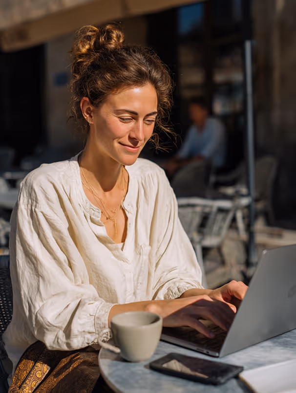 Femme souriante tapant sur un ordinateur portable à une table de café en plein air avec une tasse devant elle.