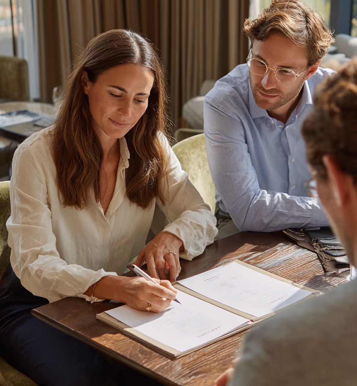 Femme signant un document avec deux hommes assis à une table en bois.