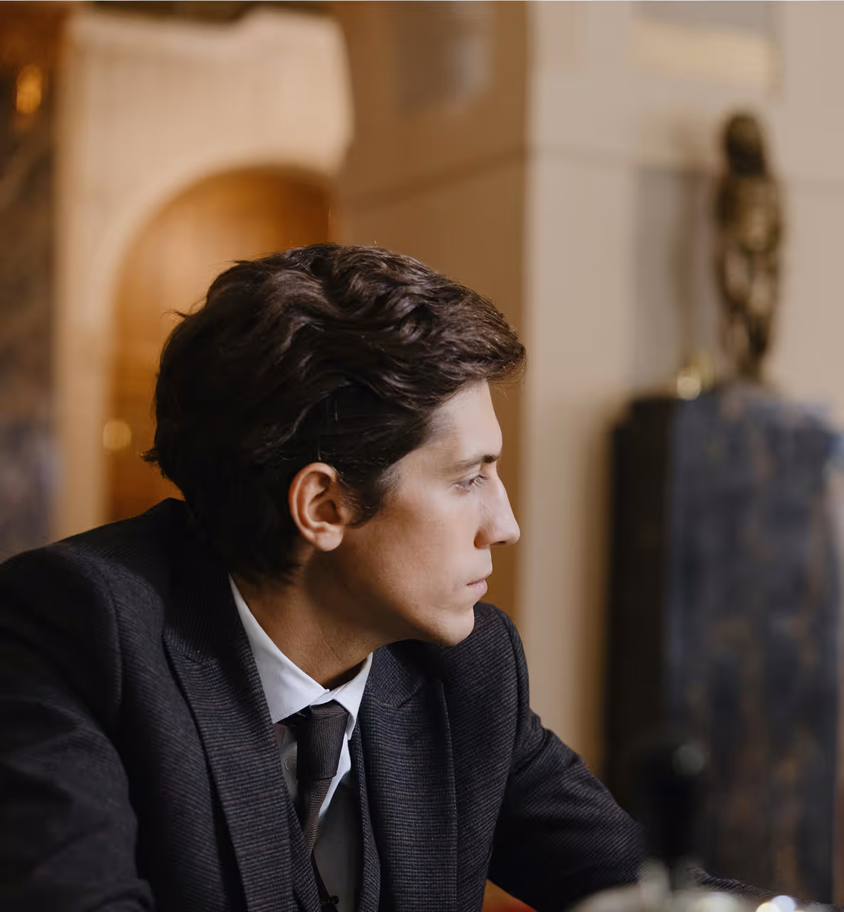 Homme en costume sombre regardant pensivement vers la droite dans un intérieur avec un mobilier classique.