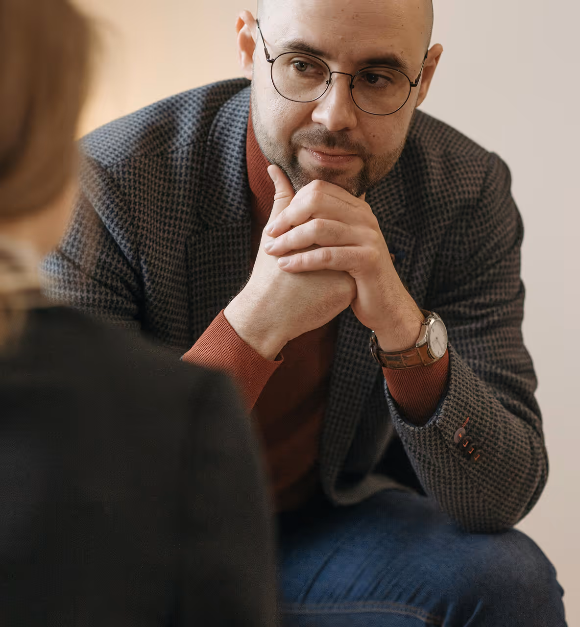Un homme chauve portant des lunettes, une veste à carreaux et un pull marron, écoute attentivement une personne non visible en avant-plan.