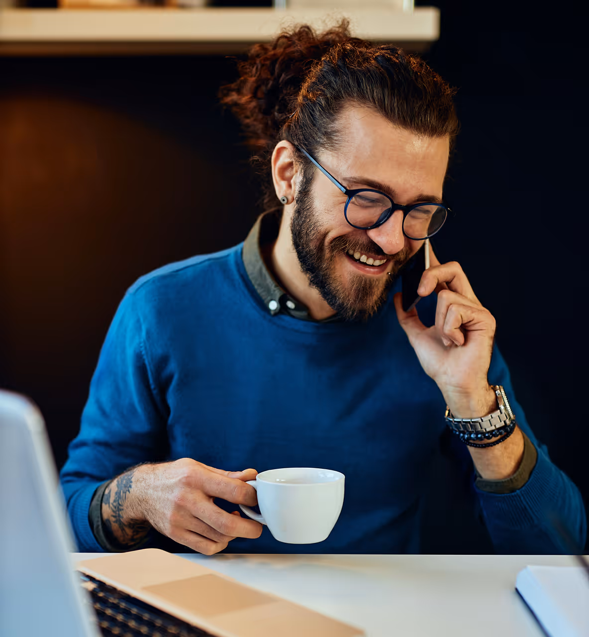 Jeune homme avec lunettes et barbe, souriant en parlant au téléphone tout en tenant une tasse de café devant un ordinateur portable.