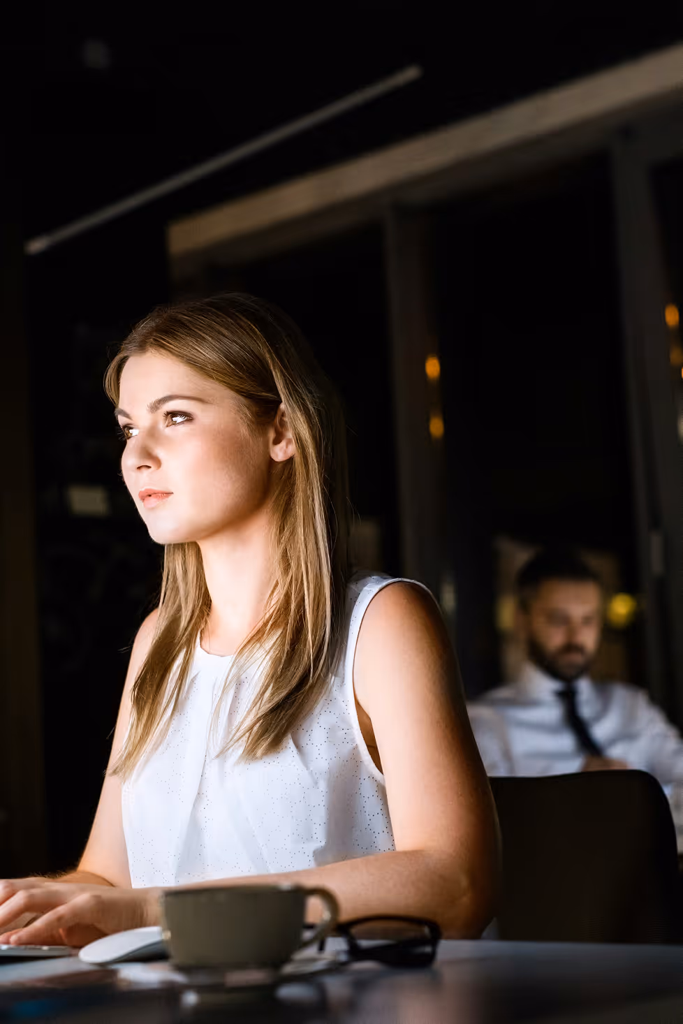 Jeune femme blonde portant un haut blanc, assise devant un ordinateur avec une tasse et des lunettes sur la table, regardant par la fenêtre, un homme en arrière-plan flou.