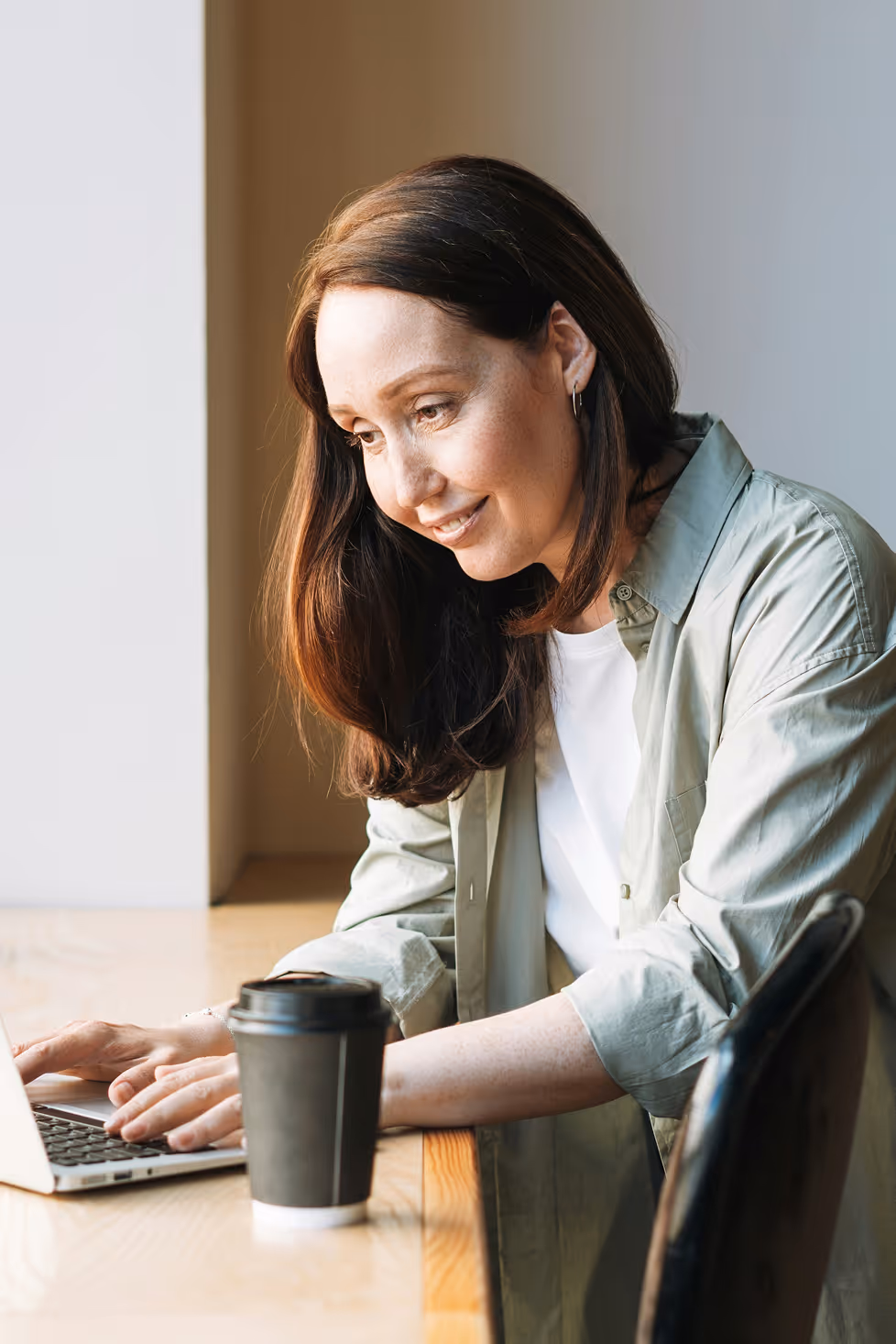 Femme souriante en chemise verte utilisant un ordinateur portable avec un gobelet de café sur une table en bois.