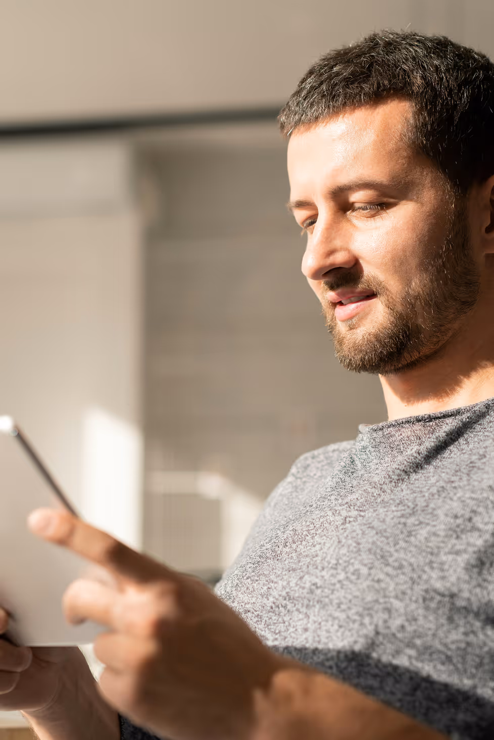 Homme souriant en pull gris utilisant une tablette tactile à la lumière du jour.