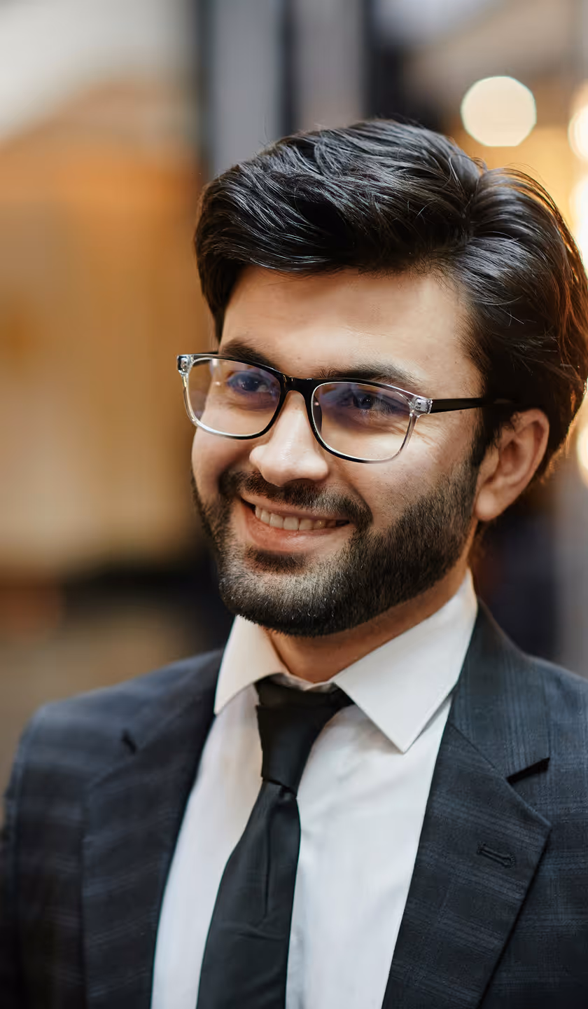 Homme souriant portant des lunettes, un costume sombre avec une chemise blanche et une cravate noire.