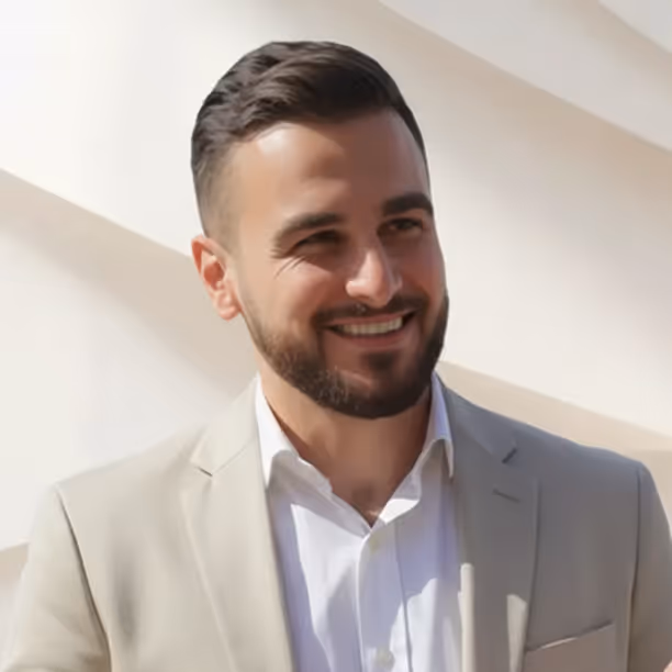 Homme souriant avec une barbe, portant un costume beige et une chemise blanche sur fond clair.