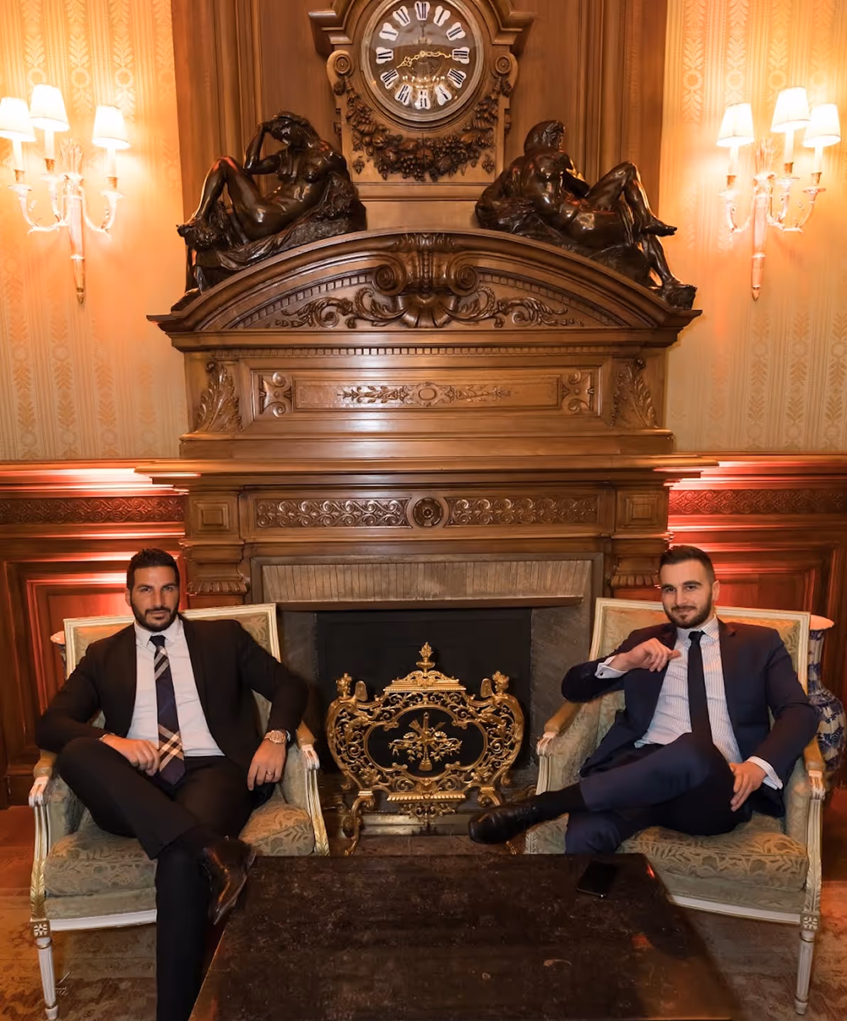 Deux hommes en costume assis sur des fauteuils verts devant une grande cheminée en bois décorée avec des sculptures et une horloge en haut.
