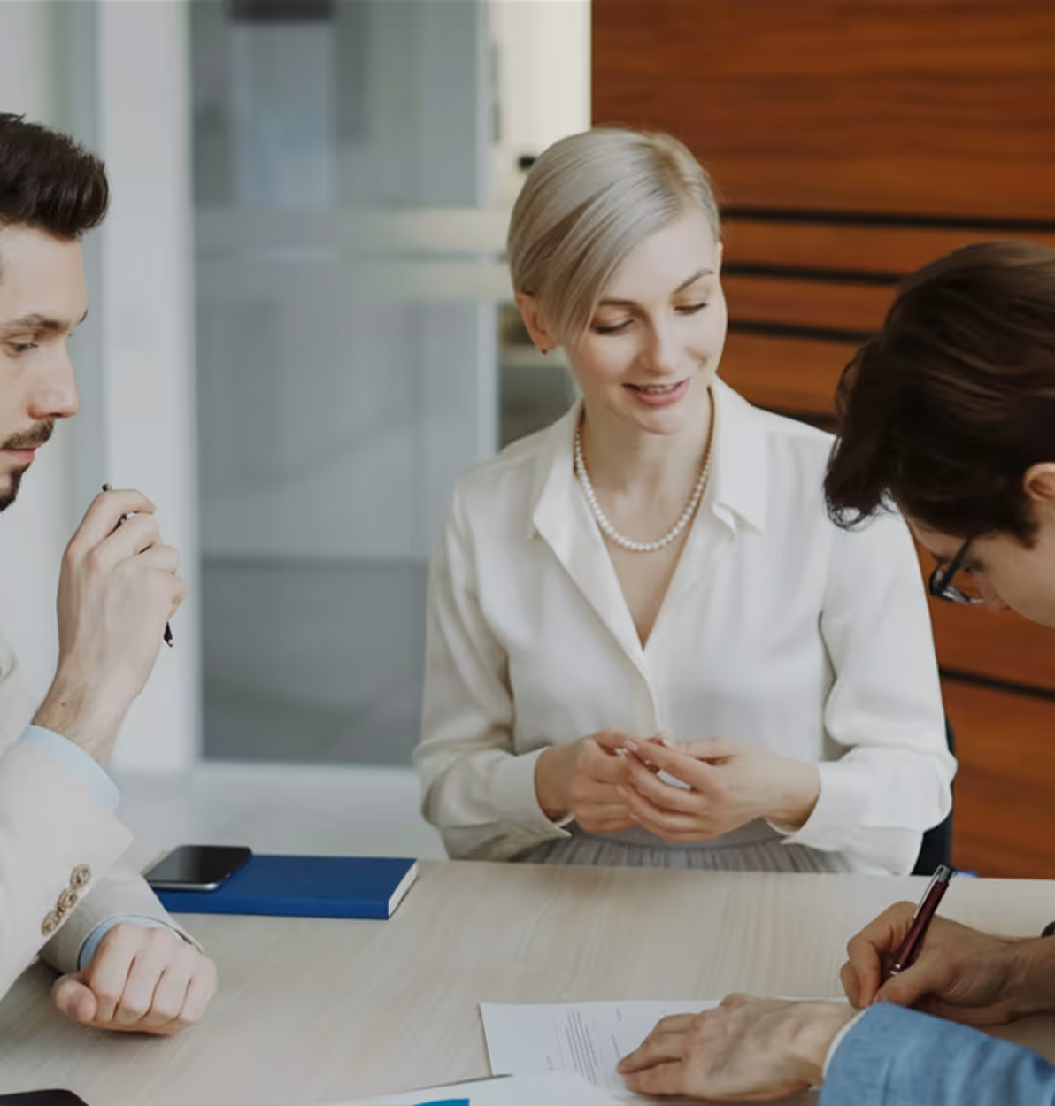 Trois professionnels discutent autour d'une table, une personne signe un document pendant que les autres observent.