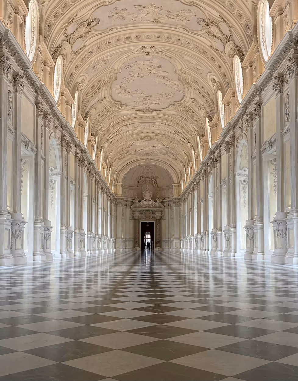 Vue intérieure d'un grand corridor baroque avec un plafond voûté orné et un sol en damier blanc et gris.