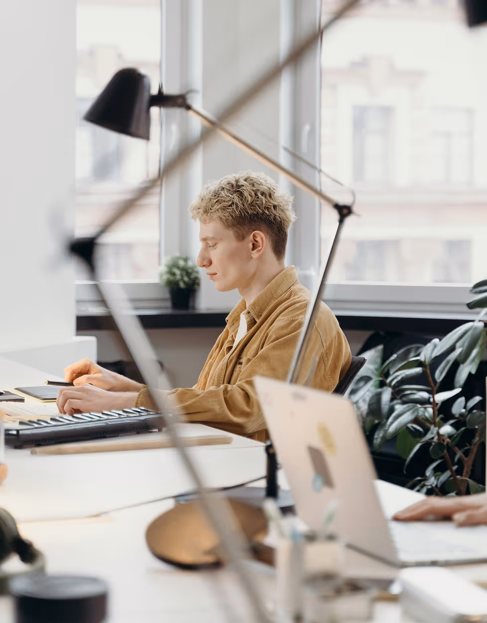 Un homme aux cheveux blonds et veste camel travaillant à un bureau dans un bureau moderne avec une lampe et un ordinateur portable.