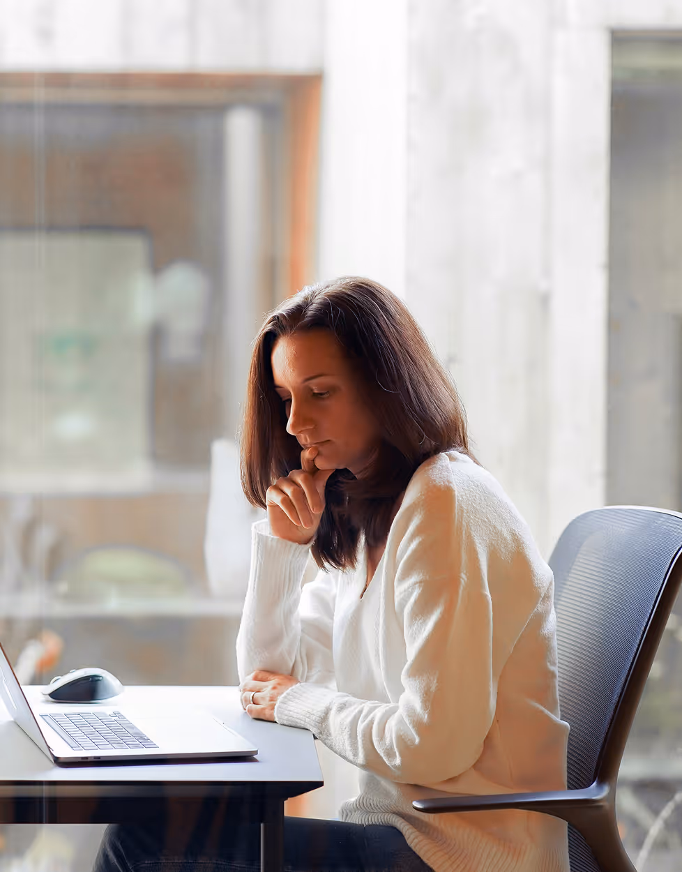 Femme réfléchissant devant un ordinateur portable dans un bureau lumineux.