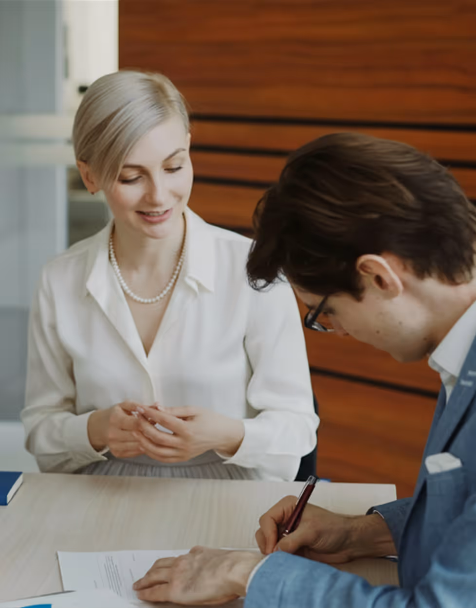 Une femme souriante en chemise blanche discute avec un homme en costume bleu qui signe un document sur une table.