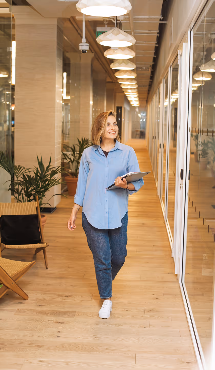 Femme souriante en chemise bleue tenant un dossier, marchant dans un couloir moderne de bureau avec des plantes et des lampes suspendues.