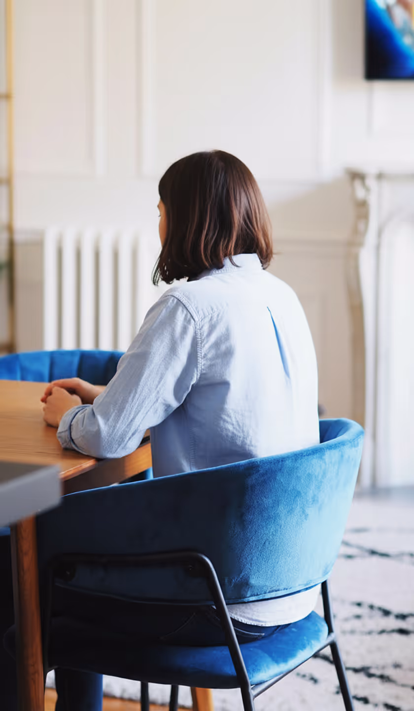 Person assise de dos sur une chaise bleue velours dans une pièce lumineuse, mains posées sur une table en bois.