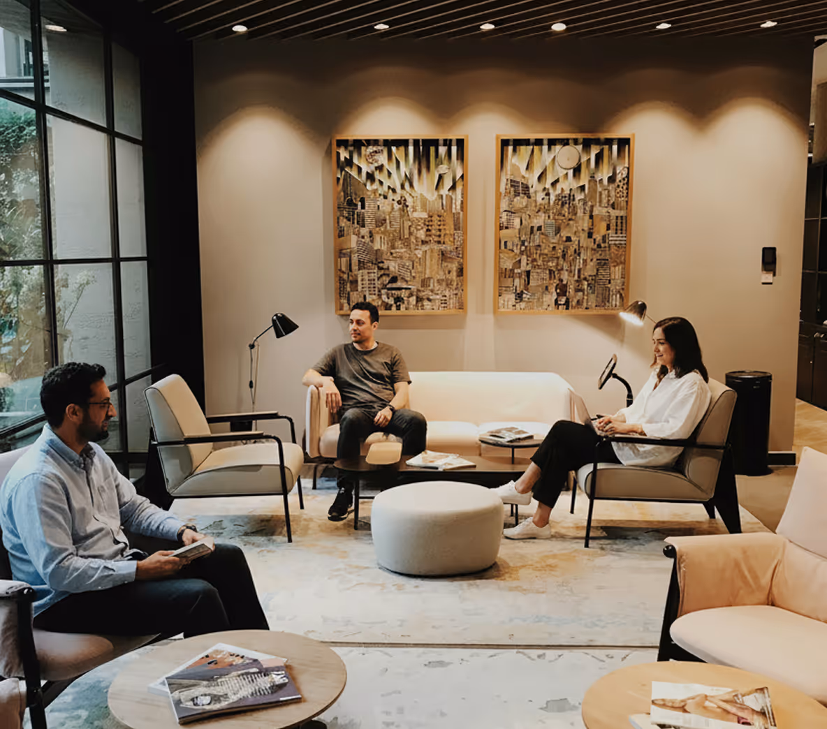 Trois personnes assises dans un salon moderne avec des fauteuils, deux tableaux abstraits au mur et une grande fenêtre sur la gauche.