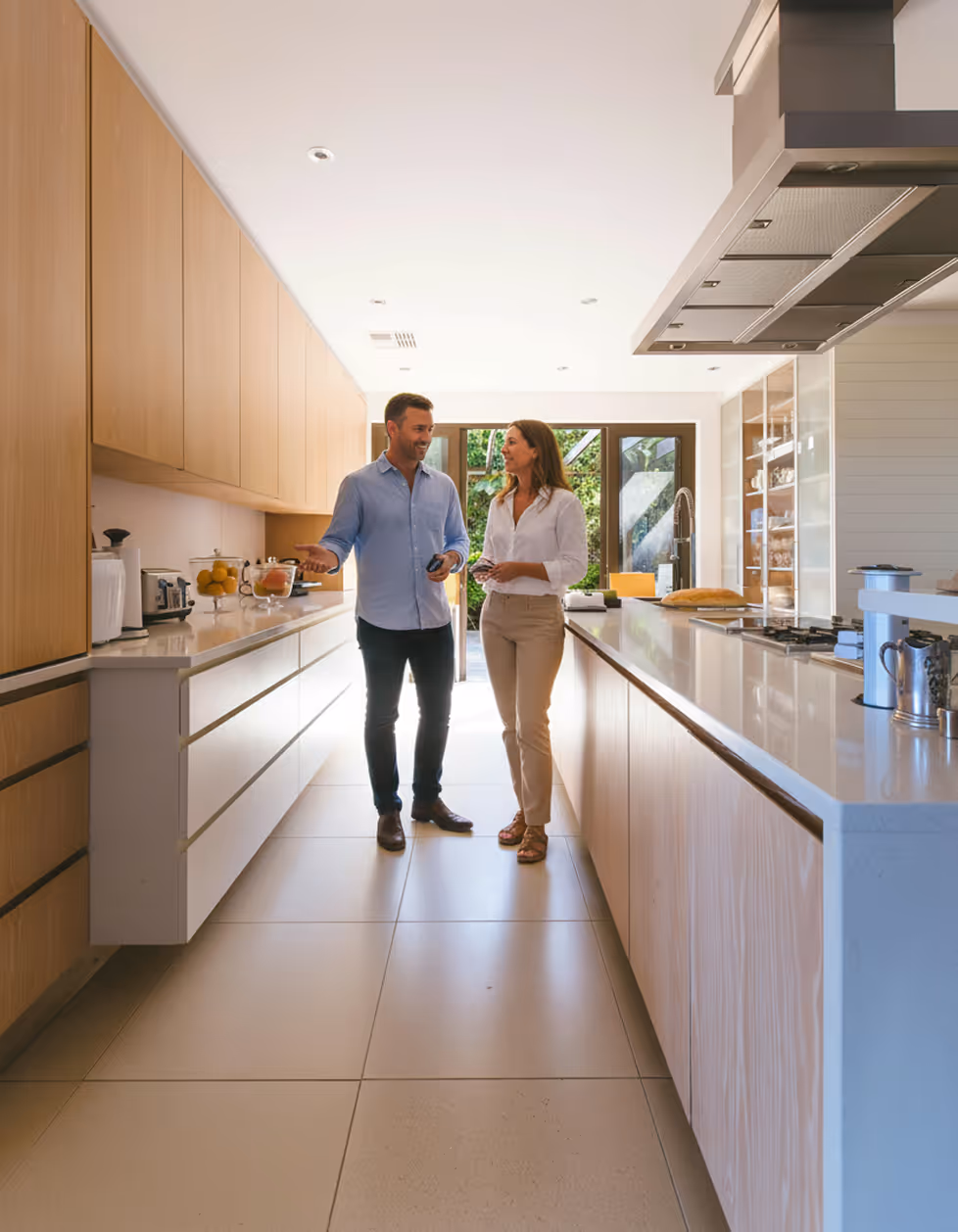 Un homme et une femme discutent dans une cuisine moderne lumineuse avec des armoires en bois clair et un îlot central blanc.