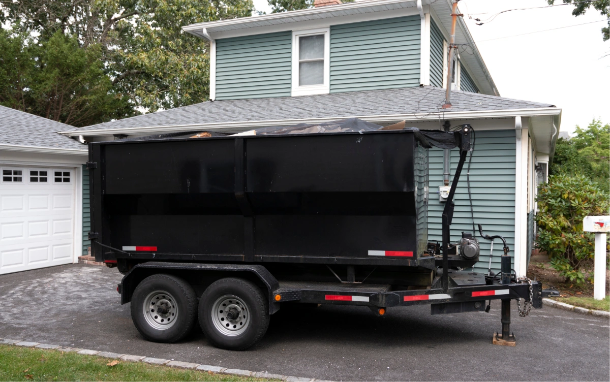 Large black dumpster container on a dual-axle trailer parked in a driveway beside a blue house.
