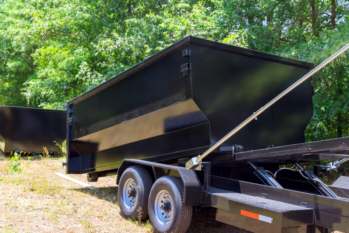 Black metal dumpster container on a dual-axle trailer parked on dry grass with trees in the background.