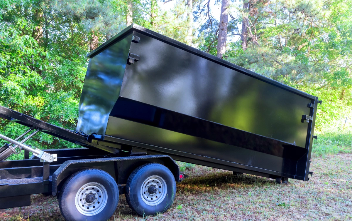 Black large metal dumpster container tilted on trailer wheels in grassy wooded area.