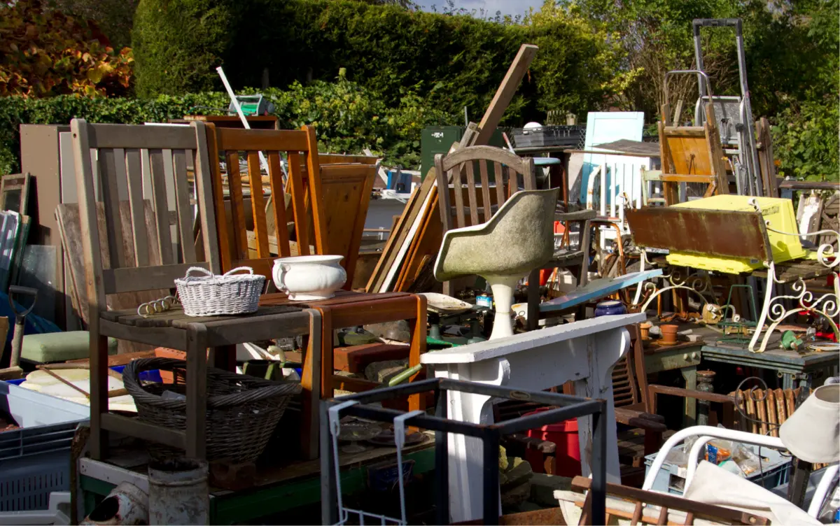 Outdoor scene showing an assortment of old and mismatched wooden chairs, baskets, and other furniture items piled together in a cluttered yard.