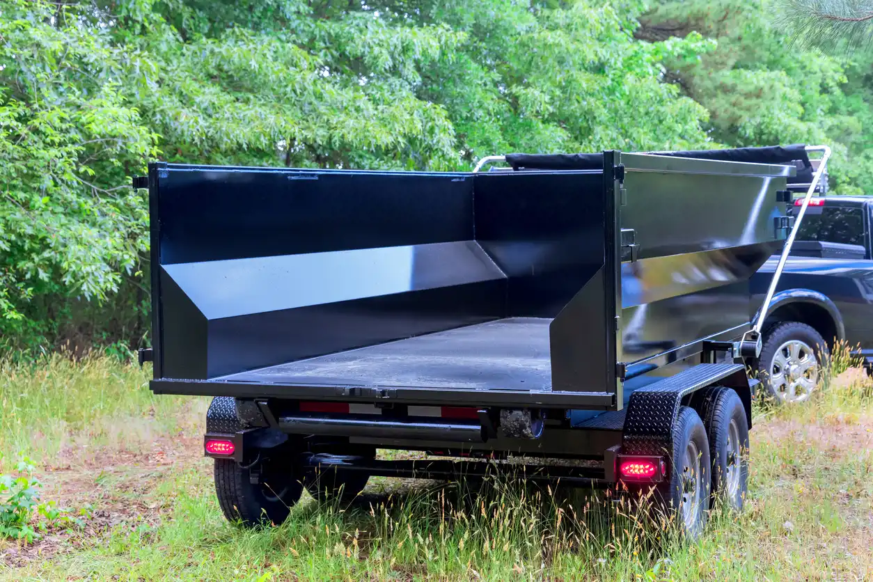 Empty black roll-off dumpster trailer with dual axles parked on grass near trees.
