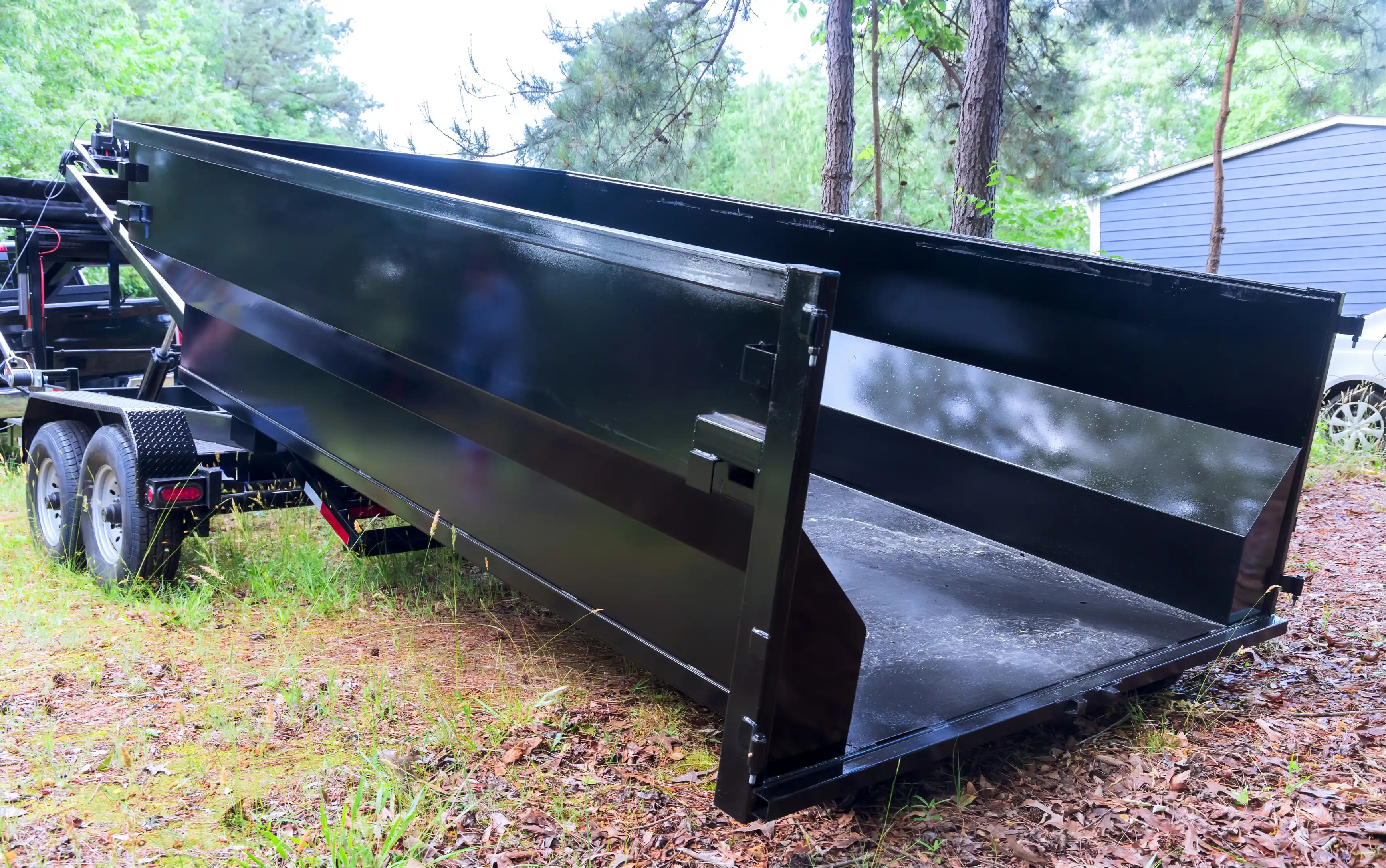 Black roll-off dumpster trailer with open back doors on grass near trees and parked cars.