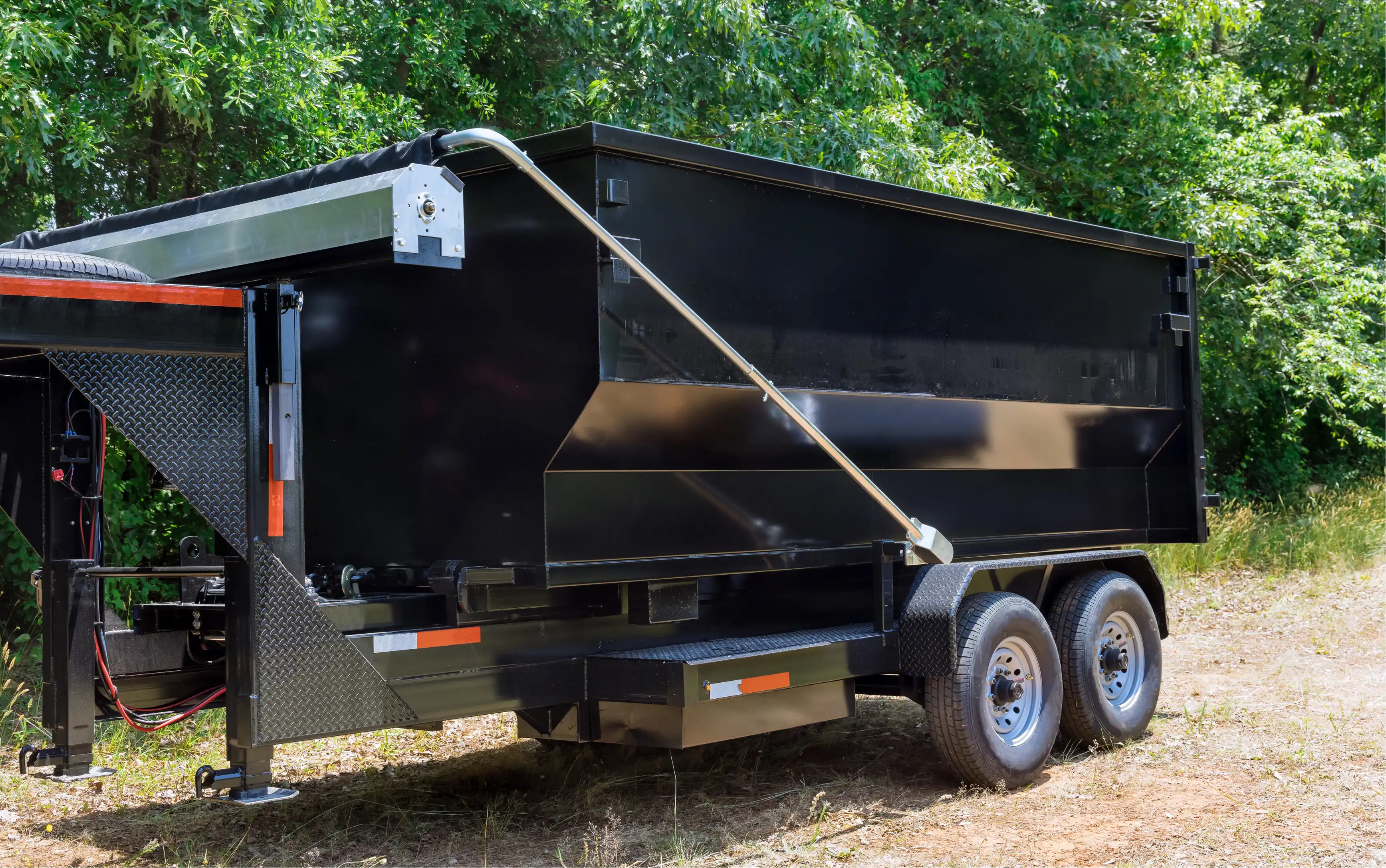Black roll-off dumpster trailer with dual axle parked on dirt near green trees.