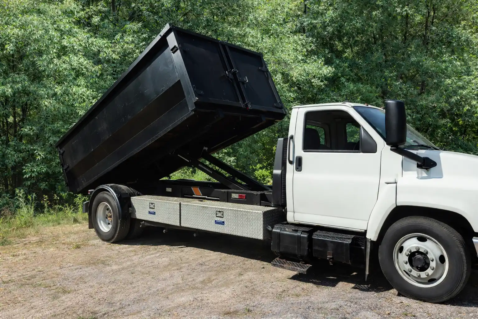 White dump truck with raised black bed parked on a dirt surface near green trees.