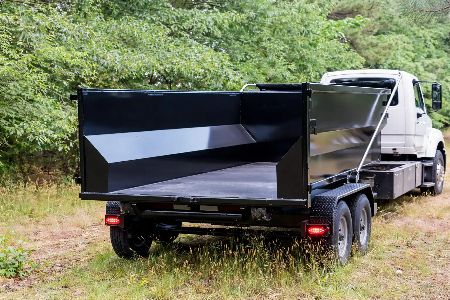 Rear view of an empty black dump truck bed attached to a white cab truck parked on grassy ground with surrounding trees.