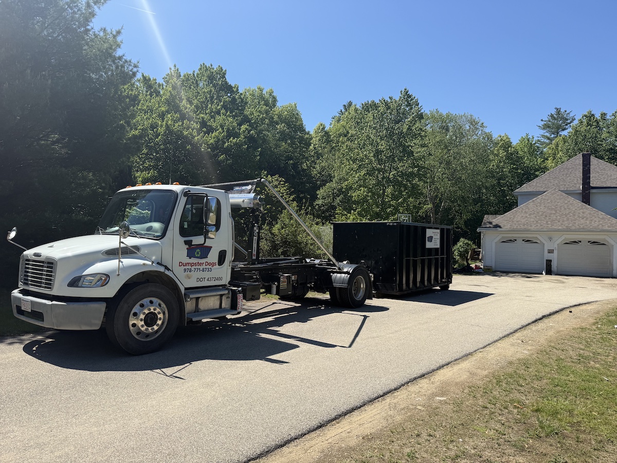 Black roll-off dumpster trailer with dual axle parked on dirt near green trees.