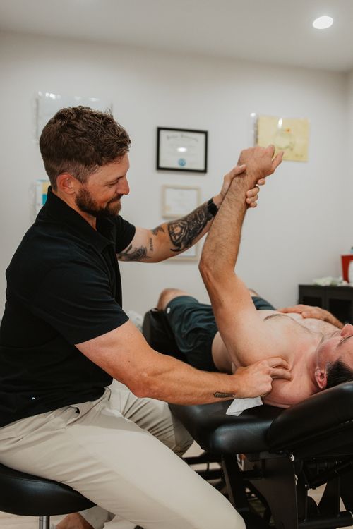 Therapist performing shoulder stretch on a shirtless man lying on a treatment table in a clinic room.
