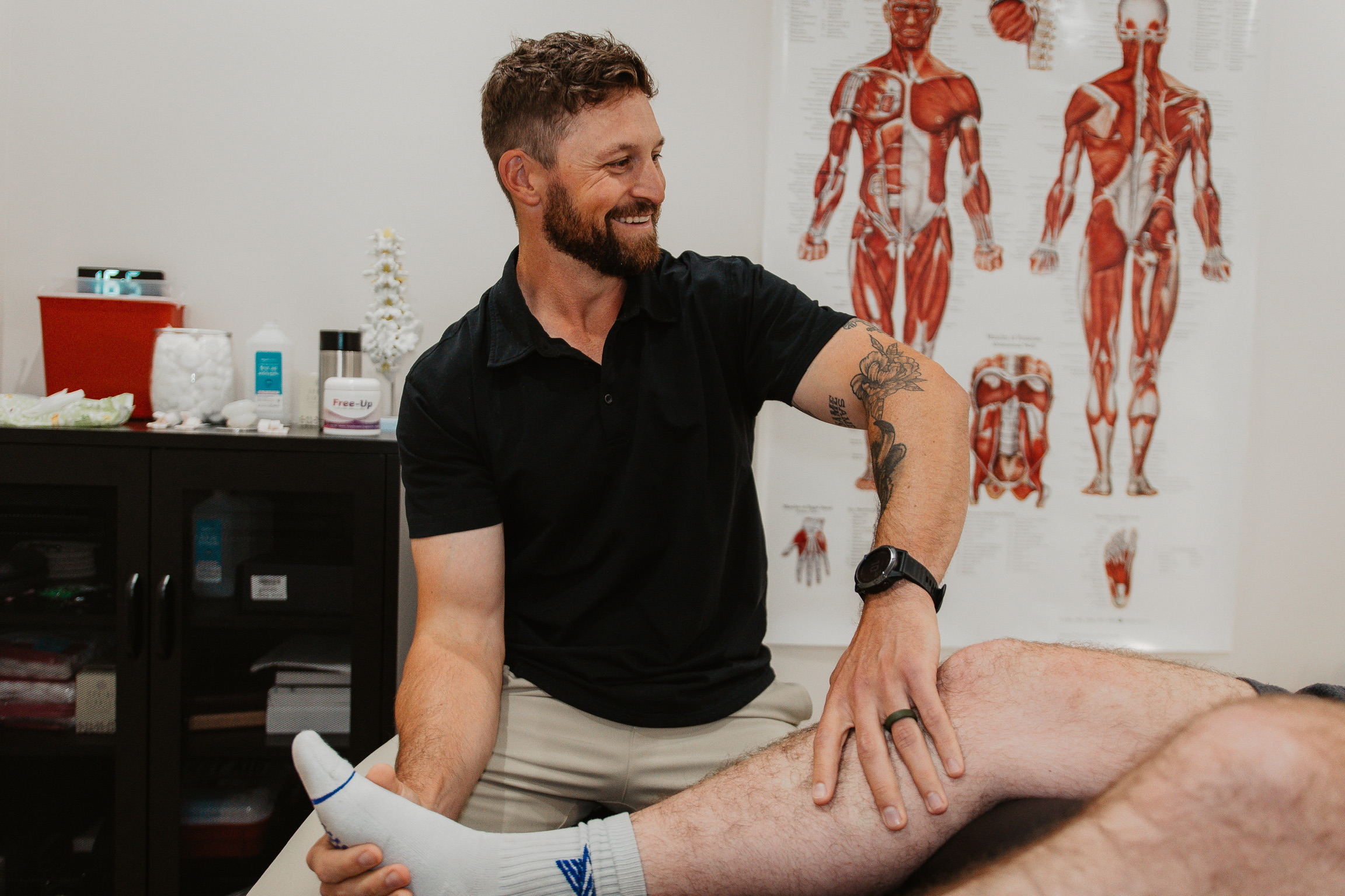 A male physical therapist with a tattoo and beard smiling while stretching a patient's leg in a clinic.