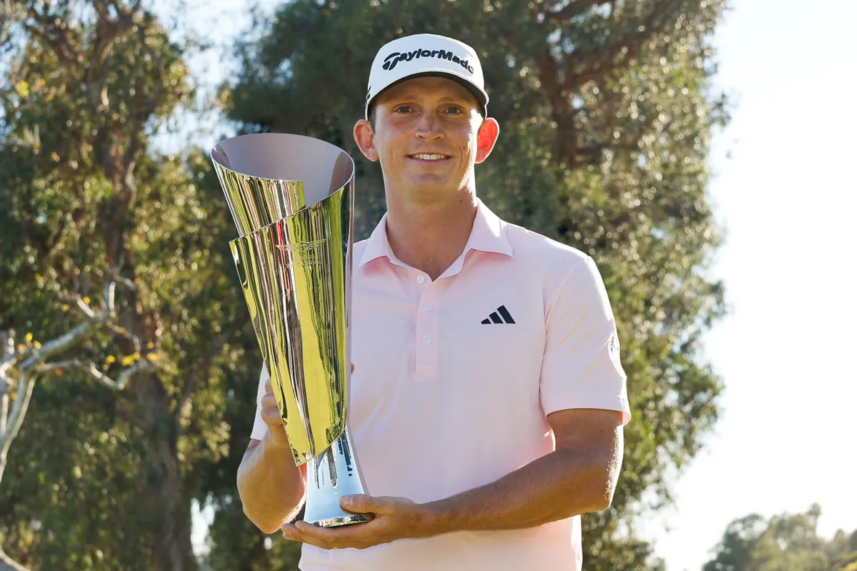 Golfer in pink shirt and white cap holding a large silver trophy outdoors with trees behind.