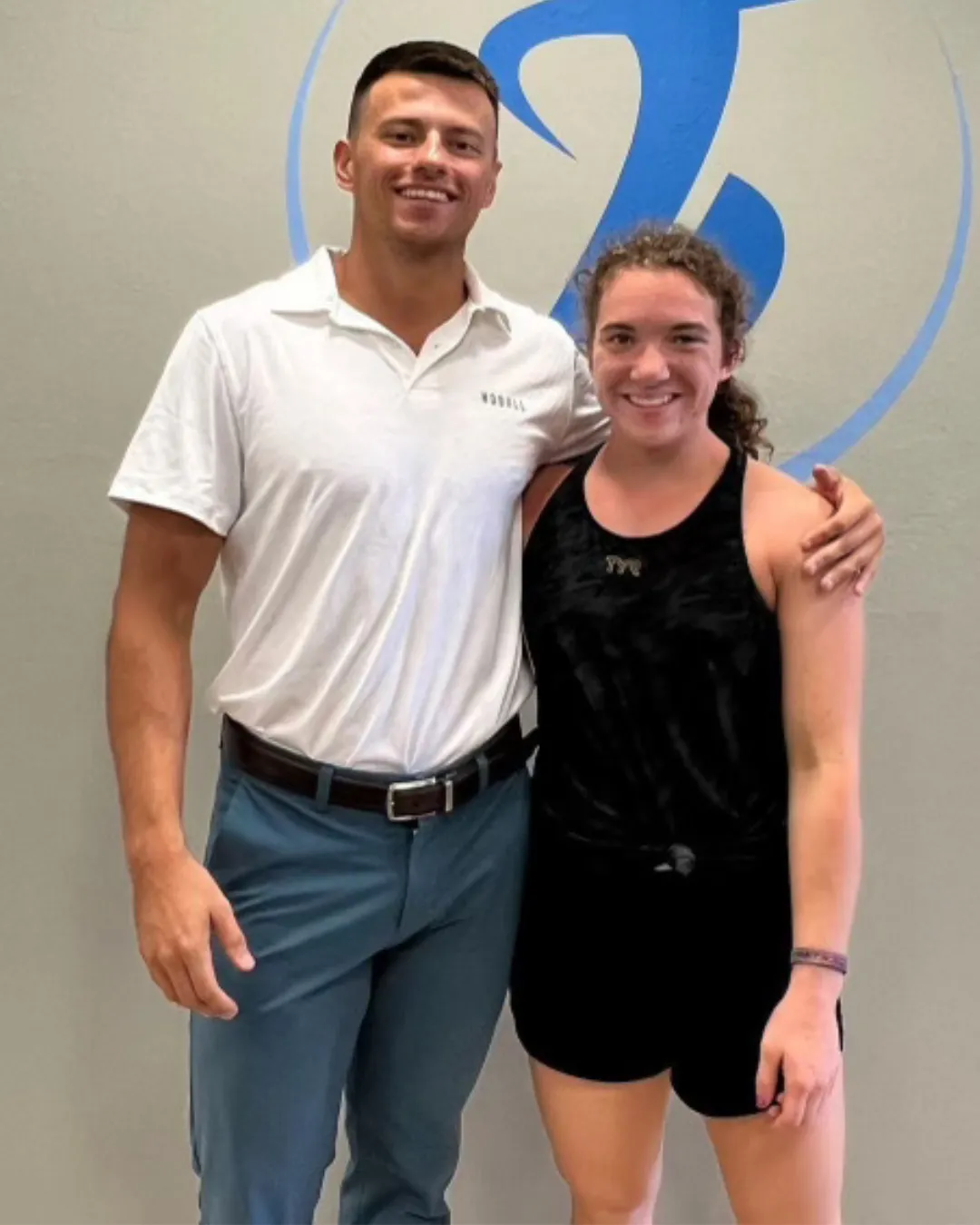 Man in white polo with arm around smiling woman in black tank top and shorts in front of a logo.