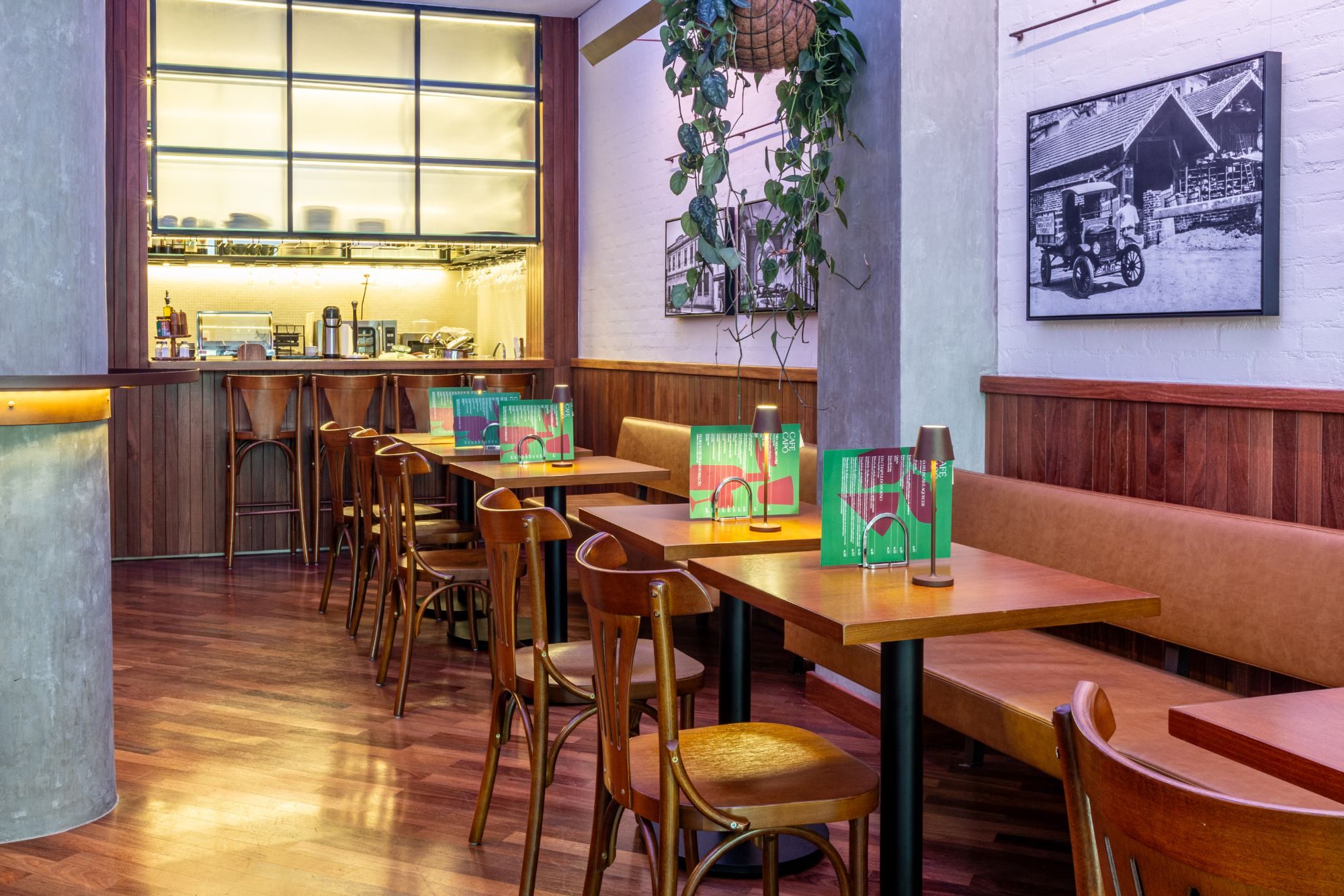 Mesas do Café Capô com luminárias e cardápios, balcão iluminado ao fundo e planta pendente