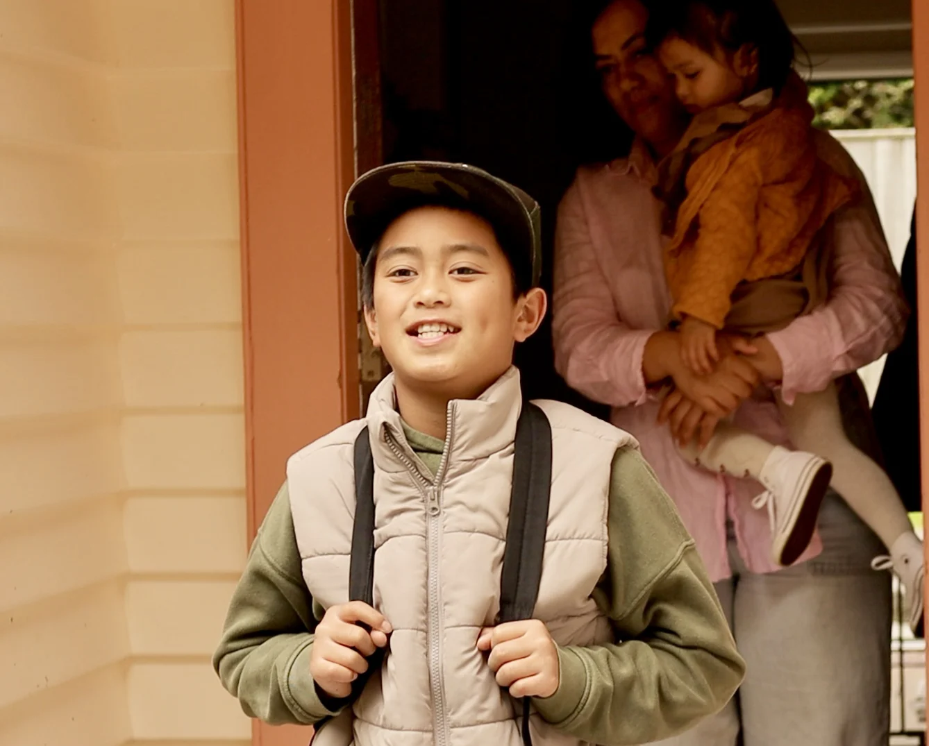 A young boy is walking out of a house, an adult walks behind him holding a toddler in her arms