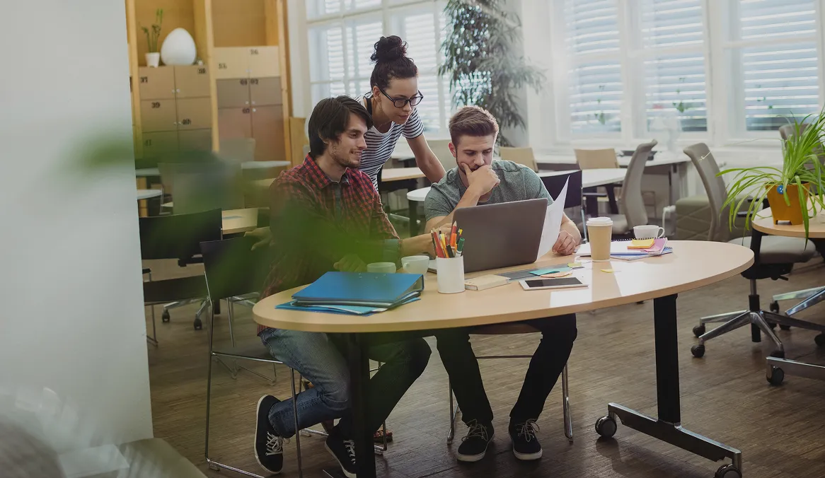 Three young coworkers collaborating at a round table with a laptop and documents in a bright office space.