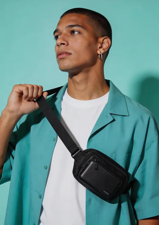 Young man wearing a teal shirt and white t-shirt holding the strap of a black crossbody bag.