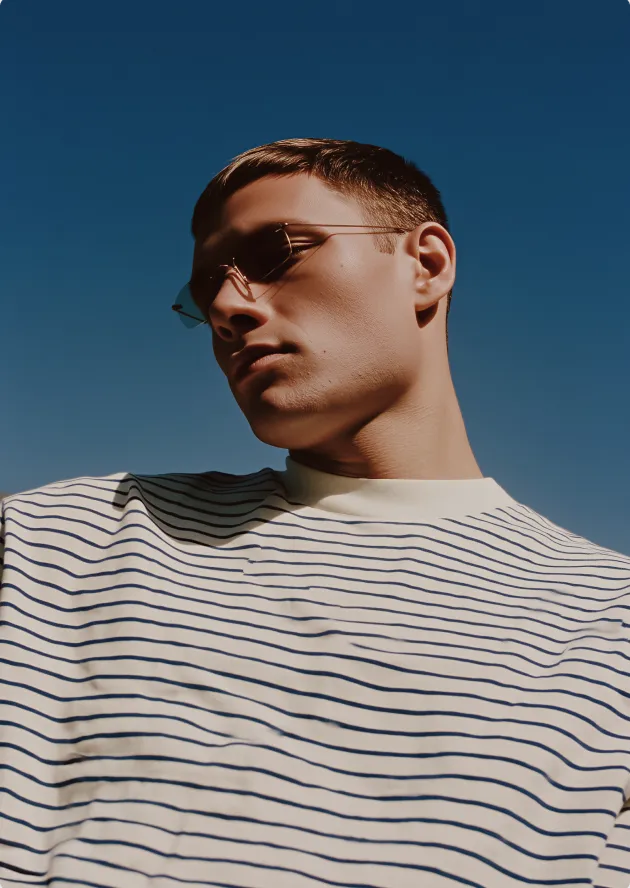 Young man wearing thin-framed sunglasses and a white shirt with navy horizontal stripes against a clear blue sky.