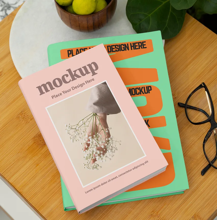 Stack of two books on a wooden table, top book with a pink cover showing a hand holding small white flowers as a mockup design.
