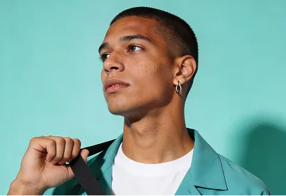 Young man in teal blazer and white shirt looking to the side while holding a black strap over his shoulder against a teal background.
