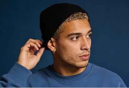 Young man with short curly hair wearing a black beanie and blue sweatshirt looking to the right against a dark blue background.