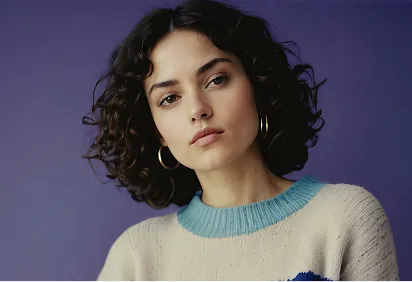 Portrait of a young woman with curly dark hair, wearing gold hoop earrings and a beige sweater with a blue collar, against a purple background.