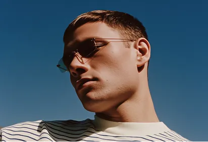 Young man wearing glasses and a striped shirt looking downward against a clear blue sky.