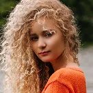 Close-up portrait of a woman with curly blonde hair wearing an orange sweater outdoors.