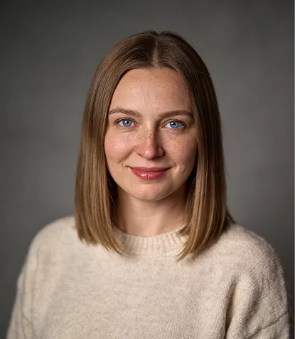 Portrait of a woman with straight light brown hair wearing a beige sweater and smiling softly against a gray background.