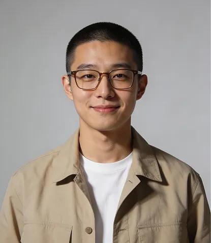 Portrait of a smiling young man with short hair, wearing glasses, a beige jacket, and a white t-shirt against a gray background.