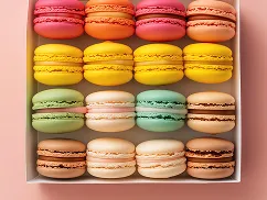Box of colorful macarons arranged in four rows with flavors in pink, orange, yellow, green, white, and brown hues.